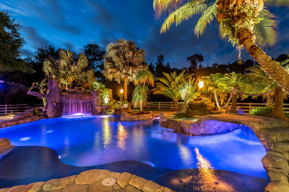 Resort-style pool at night with rock waterfall grotto, tiki torches, and tropical palms glowing in blue light